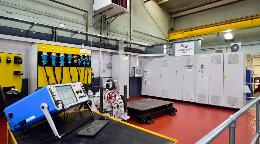 11Industrial motor test bay with condition monitoring equipment, showing a blue diagnostic console on a workbench in the foreground and large grey electrical control cabinets, cables and testing machinery in a clean workshop environment.