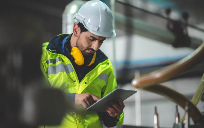 11Engineer in high-visibility jacket and hard hat using a digital tablet in an industrial setting, with yellow ear protection around their neck and blurred machinery in the foreground.